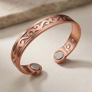 May include: A copper-colored metal cuff bracelet featuring an embossed swirling pattern on the outer surface. The interior of the open-ended band displays two circular silver-toned magnetic inserts at each terminal end for a secure fit.