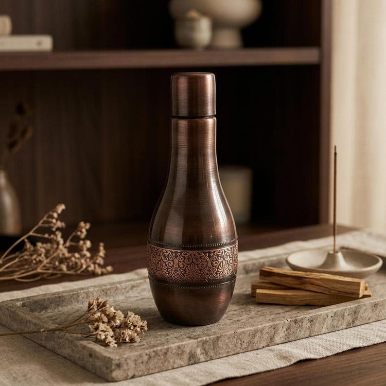 A dark copper bottle with an ornate band sits on a gray tray with dried botanicals and incense sticks.