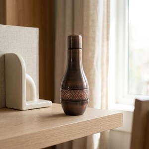 A dark copper bottle with an ornate band sits on a light wooden shelf next to a white bookend, with a window in the background.