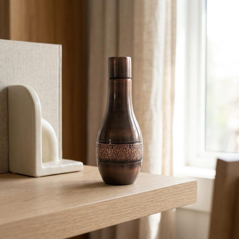 A dark copper bottle with an ornate band sits on a light wooden shelf next to a white bookend, with a window in the background.