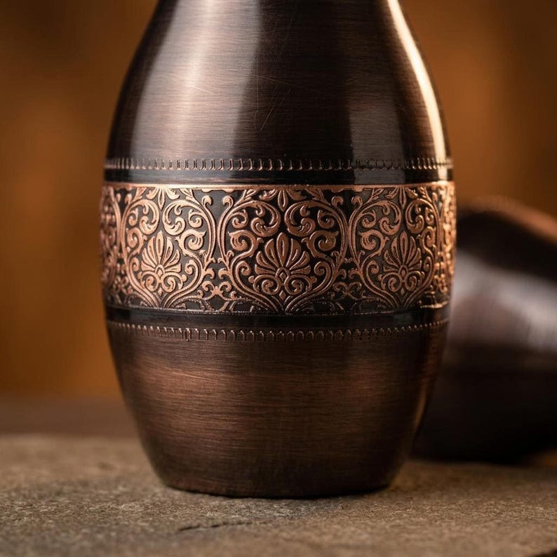 A close-up of the middle section of a dark copper bottle, showing the intricate, raised pattern of the ornate band.