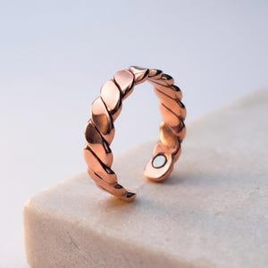 Puede incluir: Un anillo abierto en tono oro rosa con una textura trenzada similar a una cuerda a lo largo de la banda. El anillo tiene un acabado metálico pulido e incluye un pequeño imán circular incrustado en la superficie interior de un extremo.