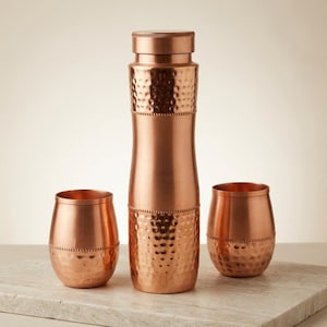 May include: A tall, cylindrical copper water bottle with a smooth center and hammered texture on the top and bottom sections, accompanied by two matching hammered copper tumblers, each featuring a decorative beaded band around the middle.