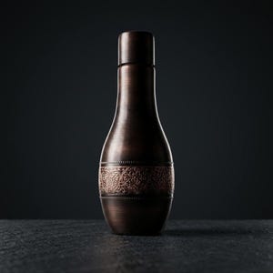 A dark, hammered copper water bottle with a narrow neck, a wide body, and a decorative band of intricate floral patterns around its center, stands upright on a dark, textured surface against a dark background.