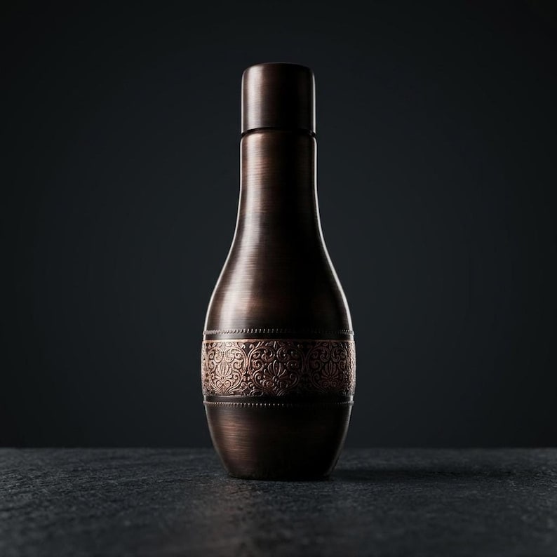 A dark, hammered copper water bottle with a narrow neck, a wide body, and a decorative band of intricate floral patterns around its center, stands upright on a dark, textured surface against a dark background.