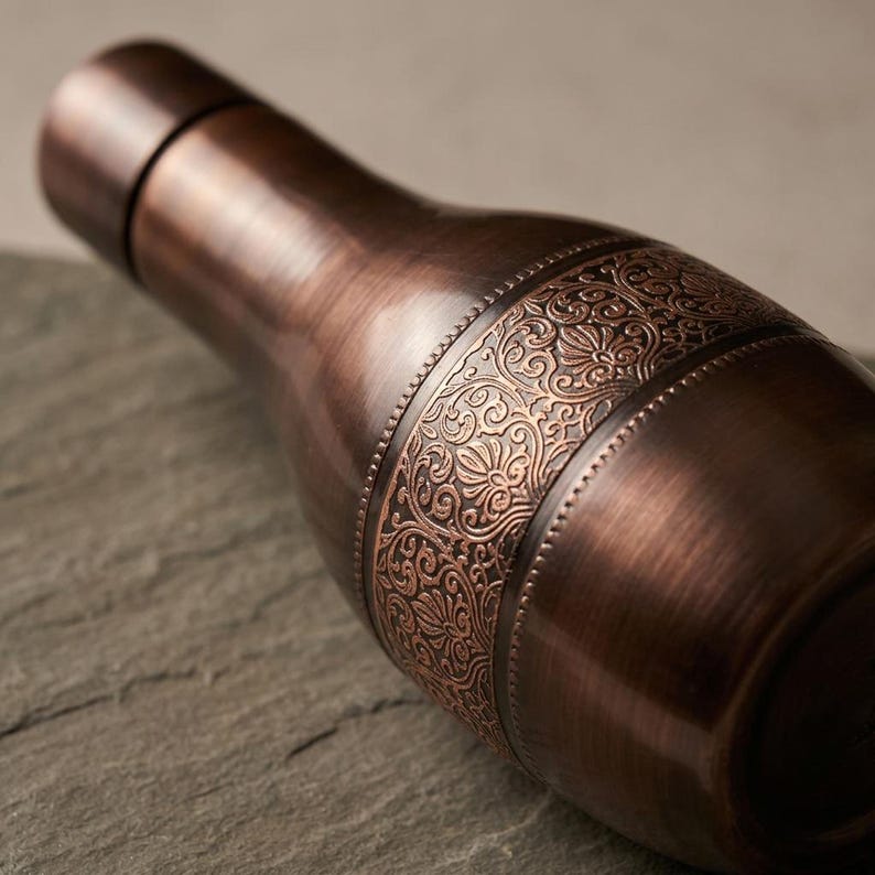 A dark copper bottle with an ornate band is angled on a dark, textured surface, showing its side and cap.