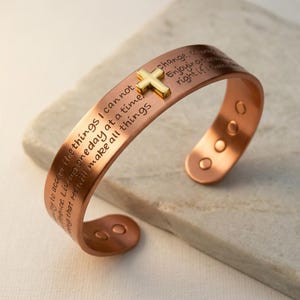 May include: A copper cuff bracelet featuring an engraved prayer text and a small gold-toned cross charm attached to the top. The open-ended band has a smooth finish with visible circular indentations on the inner ends of the metal cuff.