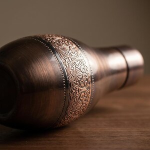 A close-up of a dark copper bottle lying on its side on a wooden surface, highlighting the ornate band.
