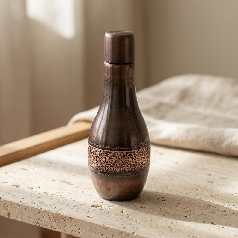 A dark copper bottle with a hammered texture and an ornate band around its middle sits on a light stone surface with a white cloth in the background.