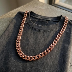 Può includere: Una collana a catena cubana color oro rosa lucido appoggiata su una t-shirt a girocollo grigio antracite scuro. La collana presenta spesse maglie metalliche intrecciate con una finitura lucida che riflette la luce sulla superficie del tessuto.