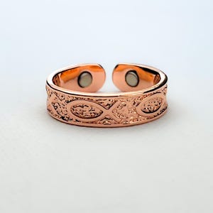 Op de afbeelding: Een open ring in roségoudkleur met een getextureerd oppervlak en herhalende vissymbolen. De binnenzijde van de ring bevat twee kleine ronde magneten die in de open uiteinden zijn verwerkt, wat zorgt voor een opvallende metalen afwerking.