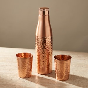 May include: A copper water bottle with a smooth neck and a hammered texture on the lower body, accompanied by two matching hammered copper tumblers. The set features a metallic finish with a warm reddish-brown tone and a cylindrical shape.