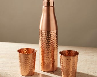 May include: A copper water bottle with a smooth neck and a hammered texture on the lower body, accompanied by two matching hammered copper tumblers. The set features a metallic finish with a warm reddish-brown tone and a cylindrical shape.