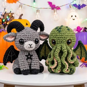 May include: Two handmade crochet plush toys. On the left, a grey goat with black horns, black wings, and a black pentagram on its chest. On the right, a green octopus-like creature with tentacles, dark green wings, and large black eyes.