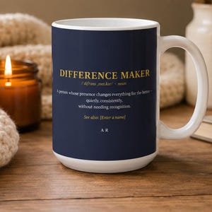 Può includere: Una tazza in ceramica blu navy scuro con interno e manico bianchi. La parte anteriore presenta un testo dorato che recita DIFFERENCE MAKER, una pronuncia fonetica, una definizione, la frase See also: [Enter a name] e le iniziali A R in basso.