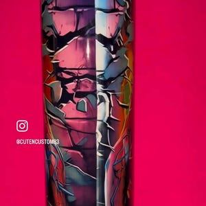 May include: A cylindrical tumbler featuring a vibrant graphic design with a cracked, shattered effect. The artwork displays a split color palette of deep purples, blues, and reds, incorporating comic book style imagery and the text CUTENCUSTOM83.