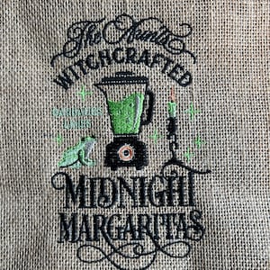 May include: A tan burlap fabric featuring black and green embroidery. The design includes a blender filled with green liquid, a frog, a candle, and the text "The Spirits WITCHCRAFTED" and "MIDNIGHT MARGARITAS" with "BARBARAS LIMES" in small print.