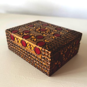 May include: A rectangular wooden box featuring a dark brown finish with a decorative pattern of small, raised circular indentations. The lid is adorned with a central design of red circular inlays surrounded by yellow heart-shaped motifs.