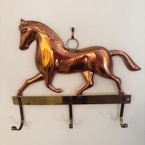 Peut inclure: Un porte-manteau mural en cuivre poli représentant une figurine de cheval en trois dimensions dans une pose de course. Le cheval est fixé à une barre horizontale en laiton avec trois crochets incurvés vers le bas et un anneau au sommet.