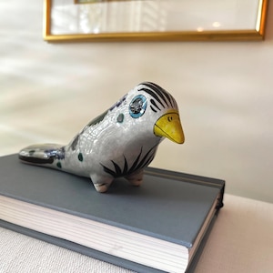 May include: A ceramic bird figurine with a glossy white finish, featuring black painted wing and tail details, a bright yellow beak, and a blue circular eye design. The bird has a rounded body shape and is positioned in a resting, forward-facing pose.