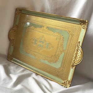 May include: A rectangular serving tray featuring a pale green centre panel framed by an ornate gold border. The tray includes two side handles with a cross-hatched pattern and a central gold embossed design with intricate scrollwork details.
