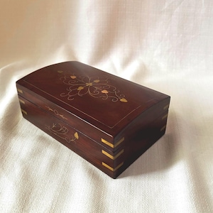 May include: A rectangular dark brown wooden box with a curved lid featuring gold-coloured metal inlay in a floral pattern. The box has brass-coloured metal corner reinforcements on the sides and a thin gold-coloured line border on the lid surface.