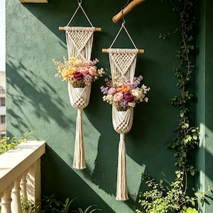 May include: Two off-white macrame wall hangings featuring woven cord patterns suspended from wooden dowels. Each piece holds a white container filled with a vibrant arrangement of artificial flowers in shades of yellow, pink, and deep red.