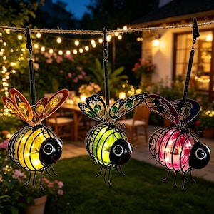 May include: Three hanging bee-shaped solar lanterns with metal wire bodies and wings. Each lantern features a glowing glass sphere in yellow, green, or pink, with black metal heads, antennae, and legs, suspended from black clips on a string.
