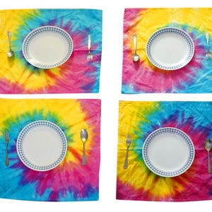 May include: Four square fabric placemats featuring a vibrant tie-dye pattern in shades of yellow, pink, and blue. Each placemat is set with a white ceramic plate with a blue checkered rim, a silver-toned metal fork, and a silver-toned metal spoon.