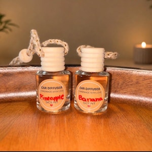 May include: Two small clear glass car diffuser bottles with light wooden caps and hanging rope cords. Each bottle features a round label with the text CAR DIFFUSER HANDMADE WITH LOVE and SCENT. One label reads Pineapple in red, the other Banana in red.