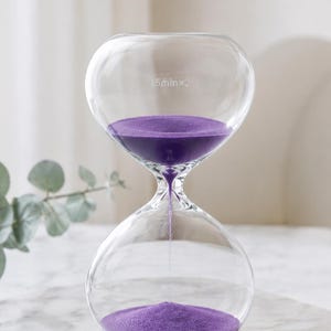 May include: A clear glass hourglass filled with vibrant purple sand. The top bulb features the white text 15minx2, indicating the duration. The sand flows through the narrow center neck into the bottom bulb, creating a functional timekeeping device.