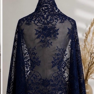May include: A dark navy blue lace shawl featuring an intricate floral pattern with scalloped edges. The sheer fabric displays a detailed botanical design woven throughout the material, draped over a mannequin to showcase the delicate texture.