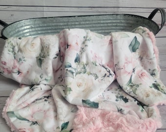 May include: A soft blanket featuring a white base with a floral pattern of pink roses and green leaves. The blanket has a textured, plush light pink border along the edges and is draped over a galvanized metal oval container with two black handles.