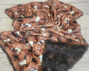Puede incluir: Una manta de felpa con fondo marrón y un patrón repetitivo de cráneos de animales blancos, lunas crecientes negras, estrellas y formas geométricas. El reverso de la manta está hecho de un material de piel sintética de color marrón oscuro.