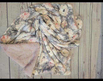 May include: A plush throw blanket featuring a floral pattern with cream, white, and soft pink flowers on a grey background. The reverse side of the blanket is a solid, textured light pink fabric with a soft, fuzzy appearance and folded edges.