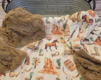 May include: A cream-colored blanket with a Western-themed pattern featuring horses, wagons, cacti, and desert landscapes. The blanket has a brown, plush border and is folded in a metal tub.