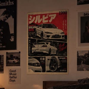 May include: A bedroom interior featuring a bed with a dark textured blanket, a black and white checkered pillow, and a round black eight-ball pillow. The wall displays a large Nissan Silvia 1999 poster, a Smiths poster, and a Blond poster.