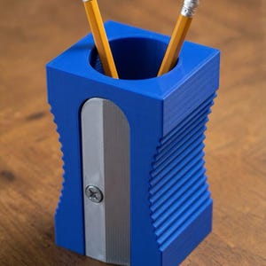 May include: A blue pencil holder shaped like a large pencil sharpener with a silver-colored front plate and a single metal screw. Two yellow pencils with pink erasers are inserted into the circular opening at the top of the blue container.