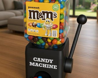 May include: A black candy machine with a clear plastic hopper filled with multi-colored peanut M&M's. The hopper features a yellow label with the M&M's logo, a character illustration, and text. A black lever is on the side, and a small tray holds loose candies.