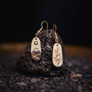 May include: A pair of dangle earrings featuring elongated, light-colored, oval-shaped pendants with gold-toned metal hooks. One pendant displays a mountain landscape with a sun, while the other features a stylized, etched line design in brown.