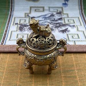 May include: A small, round, antique-style brass incense burner featuring a lid topped with a sculpted mythical creature. The vessel has two side handles, three legs, and intricate embossed floral and scrollwork patterns across its surface.