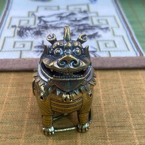 Puede incluir: Una pequeña figura de latón de estilo antiguo que representa una criatura mítica, posiblemente un león o un dragón, con detalles grabados intrincados, una boca ancha y sonriente, un cuerno prominente en la cabeza y una melena texturizada.