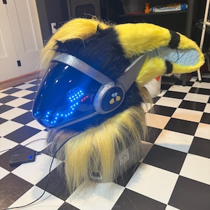 May include: A furry protogen mask featuring long, bright yellow and black faux fur, a smooth silver-coloured frame, and a dark blue visor with an integrated digital LED display showing glowing blue light patterns and a small bee icon on the side.