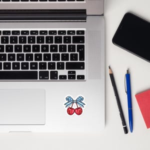 May include: A sticker featuring two red cherries with white polka dots, connected by stems to a light blue bow patterned with white stars. The sticker is placed on the silver surface of a laptop keyboard, next to a black pencil and a blue pen.