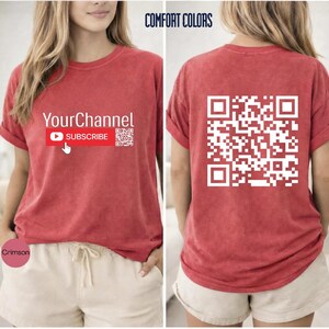 May include: A red short-sleeve t-shirt featuring the text YourChannel and a red subscribe button with a cursor icon on the front. The back of the shirt displays a large white QR code. The shirt is labeled Comfort Colors and the color is Crimson.