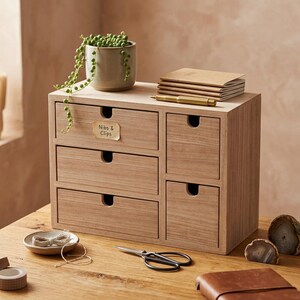 May include: A light wood desktop organizer with five drawers, featuring a label reading Nibs & Clips on the top left drawer. The unit holds a small potted plant, a stack of notebooks, and a gold pen on top, with scissors and twine nearby.