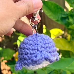 May include: A small, handmade crochet whale keychain featuring a light purple upper body and a white underbelly. The plush, textured yarn figure is attached to a silver-toned metal swivel lobster clasp and a circular split ring for easy attachment.