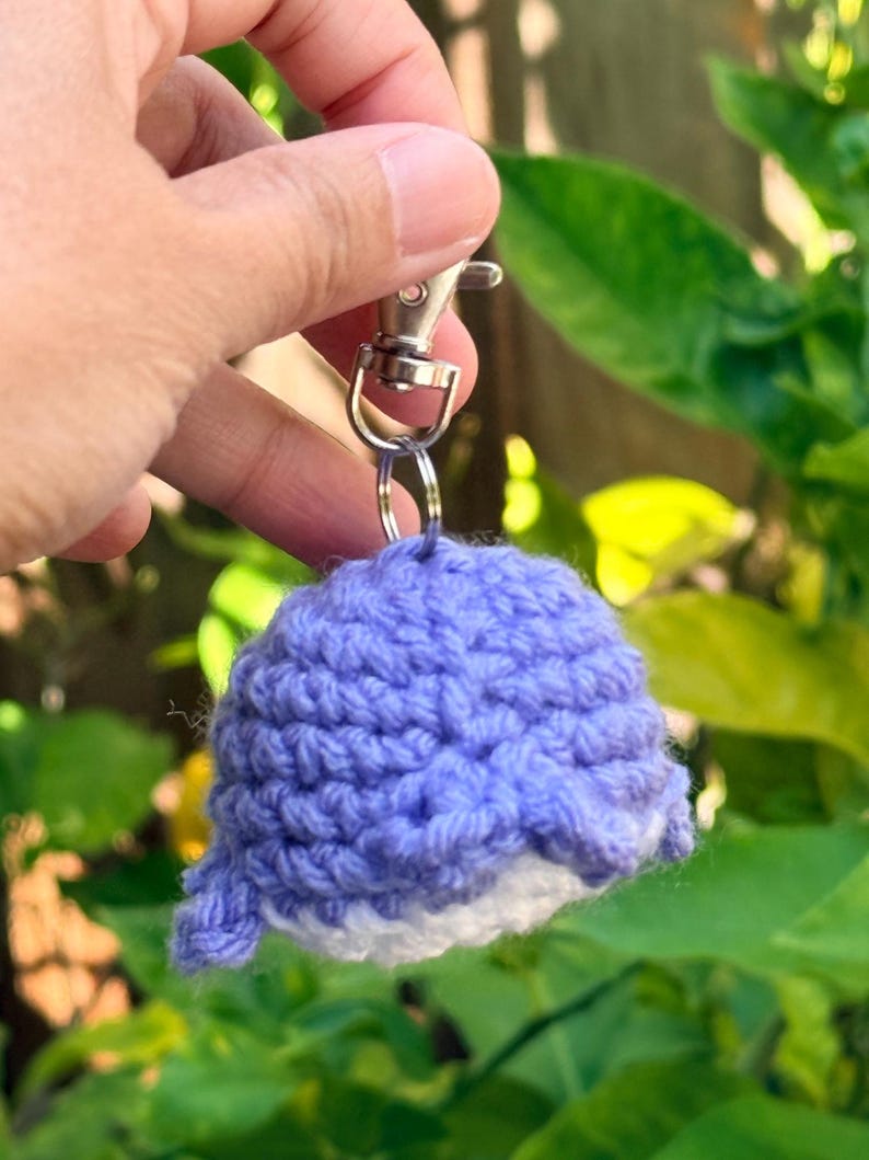 May include: A small, handmade crochet whale keychain featuring a light purple upper body and a white underbelly. The plush, textured yarn figure is attached to a silver-toned metal swivel lobster clasp and a circular split ring for easy attachment.