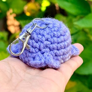 May include: A small, round, light purple crocheted plush object with a textured yarn surface. A silver-colored metal lobster claw clasp keychain is attached to the top of the crocheted item, allowing it to be easily clipped onto bags or keys.