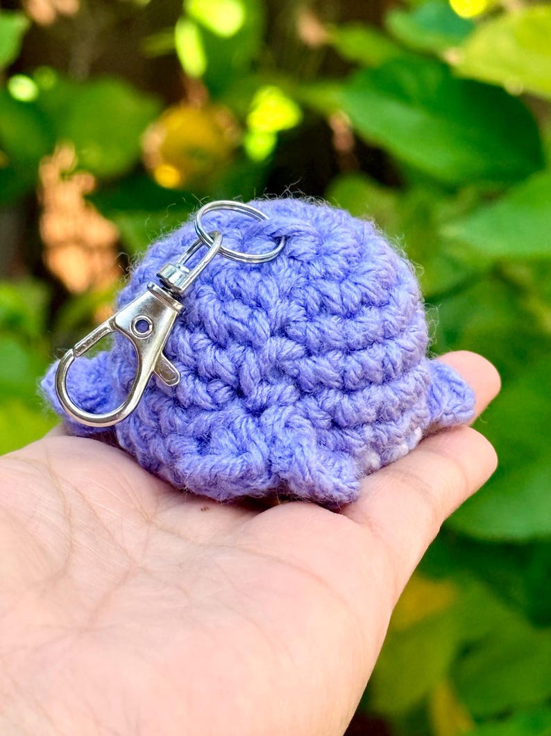 May include: A small, round, light purple crocheted plush object with a textured yarn surface. A silver-colored metal lobster claw clasp keychain is attached to the top of the crocheted item, allowing it to be easily clipped onto bags or keys.